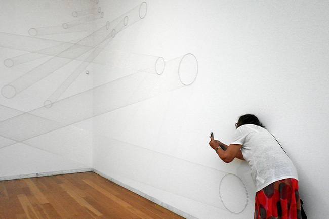 Tubos de linhas Canto de uma sala de exposição em que foram montados fios de uma parede à outra, de tal forma que, ora parecem tubos ora simples traços de um desenho. Encostada à parede direita, uma mulher tirando uma fotografia ao conjunto