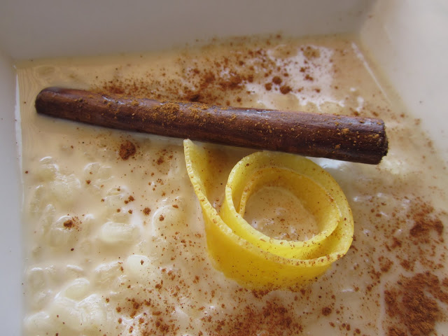 Vista panorámica de arroz con leche presentado en un plato hondo cuadrado color blanco con canela espolvoreada, además de una piel de limón haciendo un adorno en forma de espiral y una barra de canela atravesada.