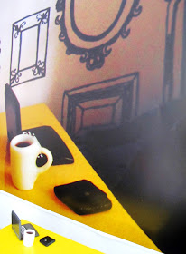 Yellow dolls' house miniature table with laptop and mug of coffee on it, in front of a blown-up photo of the same items in a dolls' house scene.