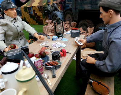 Two 1/6 scale soldiers playing cards at a table in diorama of an army post on display at a scale model exhibition.