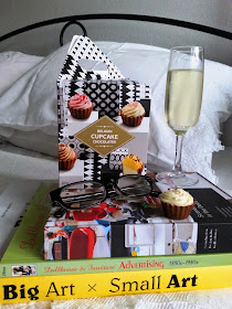 Pile of three books on a bed, with a box of miniature cupcake-shaped chocolates, a pair of reading glasses and a glass of sparkling wine on top.