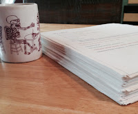 Picture Description: Stack of printed paper sits next to a coffee cup. The stack goes about half way, about an inch and a half, up the coffee cup. On the cup is a picture of a skeleton drinking coffee. Picture Description: Stack of printed paper sits next to a coffee cup. The stack goes about half way, about an inch and a half, up the coffee cup. On the cup is a picture of a skeleton drinking coffee.