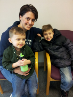 Young woman with pale skin and short, straight dark brown hair, has an arm around each of two young boys, one holding a bottle of milk.