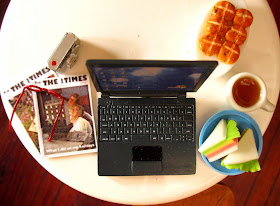 Modern dolls' house miniature round table with a laptop computer, digital camera, reading glasses, back issues of The tiny Times, sandwiches, mug of tea and hot cross buns on it.