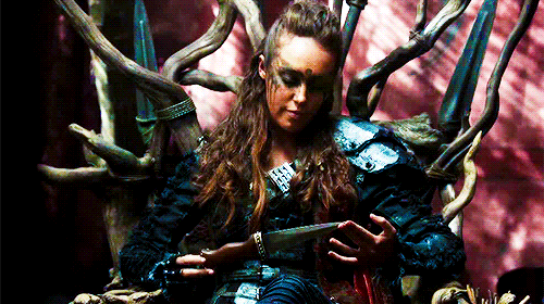 No gif: Uma garota com armadura poderosa sentada em um trono e girando uma faca na mão ameaçadoramente