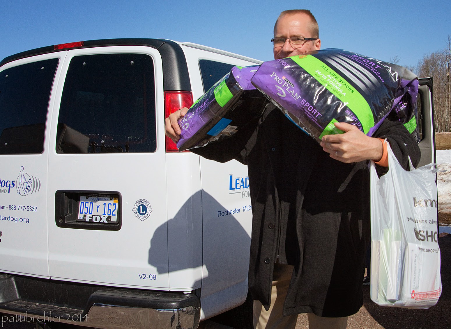 The man with khakis and a dark jacekt is carrying two large bags of Purina dog food on each shoulder, walking away from the back of a white Leader Dogs for the Blind van. It is very sunny with a blue sky.