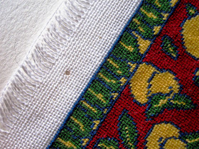 Edge of a piece of cross-stitch fabric with a dolls' house miniature rug stitched on it, showing pin marks from blocking.