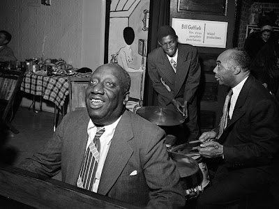 James P Johnson, smiling, playing the piano with other musicians in the background
