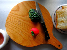 Modern dolls' house miniature chopping board with a knife, an avocado and a tomato on it. Next to it is a plate of toast and a mug of tea.