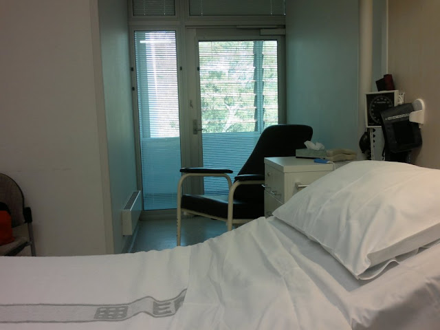 View of hospital room showing bed in the foreground, behind which is an alcove with an armchair and a glass door to a balcony outside.