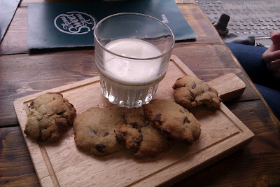 A small chopping board topped with 5 hot and freshly baked choc chip cookies accompanied by an ice cold glass of milk.
