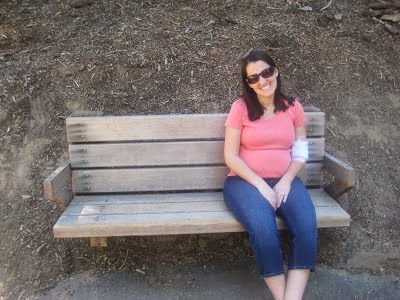 Pregnant Rachel sitting on a park bench wearing an orange shirt and sunglasses