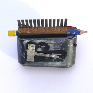 Reverse of a office finds sculpture, which uses a flattened pen clip as a fixture with which to attach it to the body.