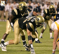 Three defensive University of Central Florida football players on the natural turf of a night football game preparing for the ball to be snapped.