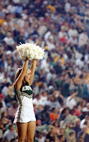 Cheerleader in green and white uniform lifts white pom poms above her head while a stadium of fans cheer on.