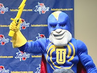 Superhero looking mascot wears a muscular blue outfit with a red cape and a yellow lighting bolt.