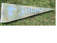 Tattered triangle shaped pennant flag with worn blue calligraphy font reading Tulane and similar colored image of a football player carrying a ball. The pennant is laid on green grass.