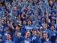A sea of University of Memphis fans in blue t-shirts in the stands at a basketball game waiving blue poms.