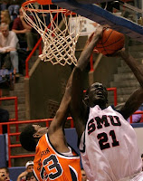 Basketball player in white uniform with blue writing reading SMU #21 takes the ball to the basket against a defender in an orange uniform.