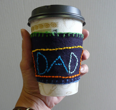 A coffee cup that is foam with a black lid with a blue cup holder with green and light blue stitching around the boarder with dad stitched in blue, with a yellow and orange border.