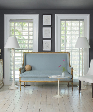 Living room with dark grey walls, white windows with white shades, natural wood floor and a blue settee with gold trim, on each side of the settee are white floor lamps and a small white tulip tray