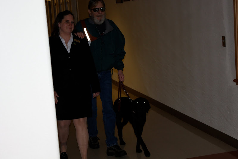 Mike walks down the hall with his hand on an instructor's shoulder heeling Millie at his side