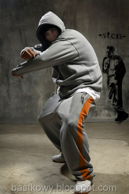 "Młody mężczyzna w szarym dresie tańczy w dynamicznej pozycji, na ścianie za nim widnieje szablon graffiti.