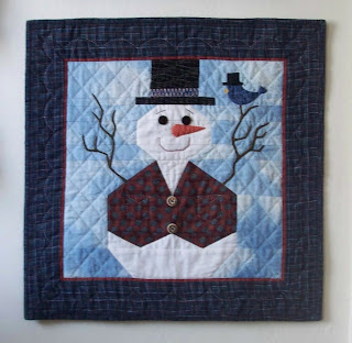 quilted wallhanging of a snowman wearing a tophat and two button plaid vest. His arms are made from twigs and a bluebird, also wearing a tophat is resting on one of the arms