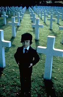 O pequeno Damien usando terno, gravata e chapéu/boina pretos, com as mãos nos bolsos em pé em um cemitério padronizado (um gramado cheio de cruzes lisas brancas quase da altura do garoto)