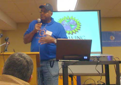 Will Allen in cap and blue sweatshirt, speaking in front of a screen with the Growing Power logo on it
