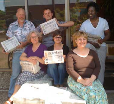 Three men and three women perched on top of a pile of newpapers, some holding small bundles of the issue