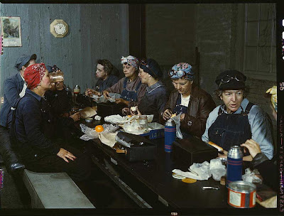 Women gathered around a long table, wearing overalls and headscarves, their lunch buckets on the table before them