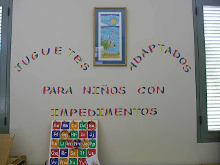 Wall in a children's classroom. Text in spanish reads: Toys adapted for children with impairments. An alphabet toy leans against the wall.
