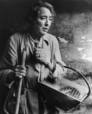 black and white photograph of Vita Sackville-West in her later years, posed with gardening basket and tools. Image source cited below.