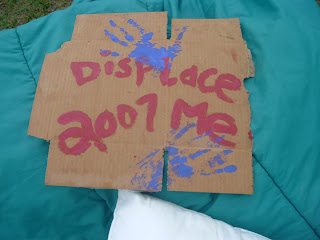 Piece of cardboard laying on a sleeping bag. The cardboard has two blue paint hand prints on it and the words ''Displace Me 2007'' in red paint