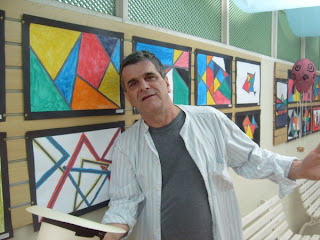 Imagem de Antonio Peticov, de camisa listrada aberta, deixando aparente a camiseta cinza, segura um chapéu de palha na mão. Ao fundo, suas telas.