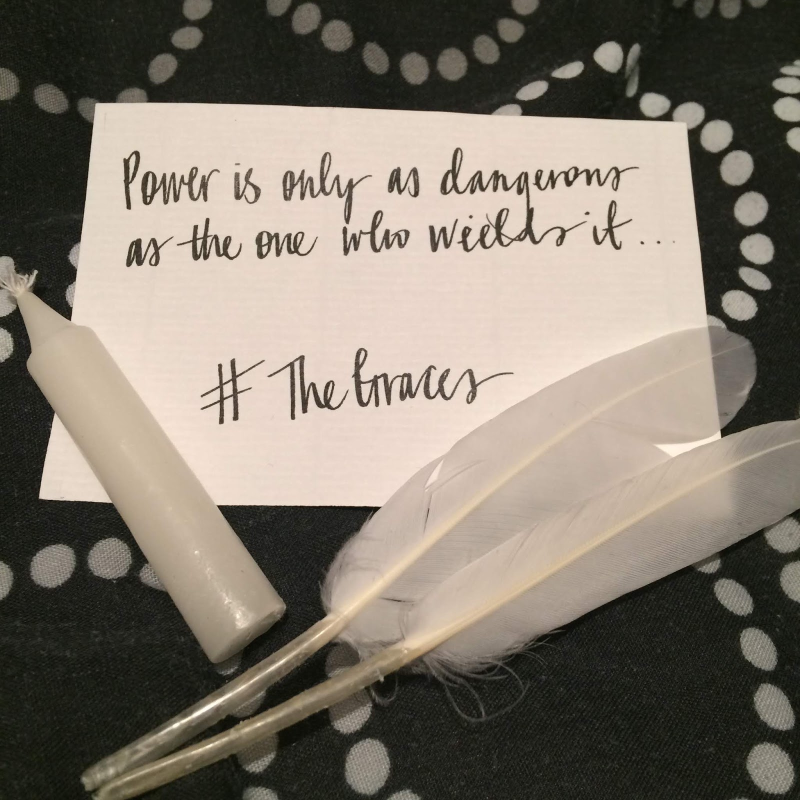 Swag for The Graces, featuring two slim, white feathers, and a slim, short, white candle, and a note saying power is only as dangerous as the one who yields it.