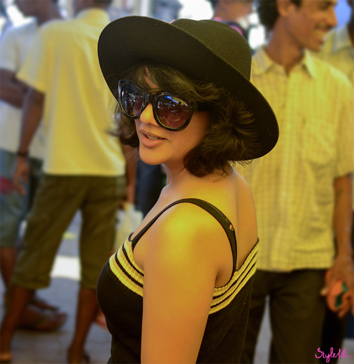An image of a woman walking through a crowded market place candidly looking back as she wears sunglasses and a hat over her short hair on a summer holiday break in Goa, India