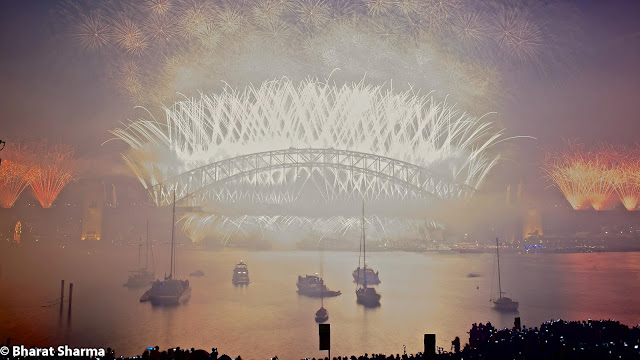 It was New Year, when Bharat and Ishita flew to Australia from New Zealand for celebrations. This Photo Journey shares some of the moments from Sydney Harbour, where some amazing fireworks were captured on New Year Evening. Let's check out more from Sydney Harbour.Above photograph shows Sydney Harbour Bridge which is basically a steel bridge through arch bridge across Sydney Harbour that carries rail, vehicular, bicycle and pedestrian traffic between the Sydney central business district (CBD) and the North Shore. The dramatic view of the bridge, the harbour, and the nearby Sydney Opera House is an iconic image of Sydney, New South Wales, and Australia. The bridge is nicknamed 'The Coathanger' because of its arch-based design.In midnight, Australia celebrates the start of a New Year! Sydney does this in style by lighting up the night sky with a spectacular fireworks display launched from seven barges on the Harbour, the rooftops of seven city skyscrapers and, unforgettably, from Sydney Harbour Bridge. It becomes a  reason why Sydney is the New Year’s Eve Capital of the World.Many of the folks come to Australia for New Year Celebrations. Many folks don't get right place at Sydney Harbour foreshore to watch what is clearly the best New Year’s Eve fireworks display on Earth. This firework shows is quite popular and there are some online services providers who stream live view from a great place and these models work very well.Dawes Point, Garden Island, Sydney Cove and Vaucluse are some of the main places at Sydney Harbour. Few helicopter cameras keep capturing the great moments for online folks. The bridge's design was influenced by the Hell Gate Bridge in New York. It was the world's widest long-span bridge, at 48.8 meters wide, until construction of the new Port Mann Bridge in Vancouver. It is also the fifth longest spanning-arch bridge in the world, and it is the tallest steel arch bridge, measuring 134 metres from top to water level. Until 1967 the Harbour Bridge was Sydney's tallest structure.