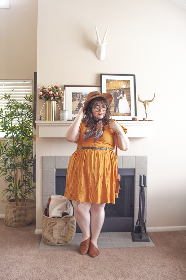 An outfit consisting of a camel brown panama hat, a brick red patterned neck scarf, a mustardy yellow skater dress and brown d'orsay flats.