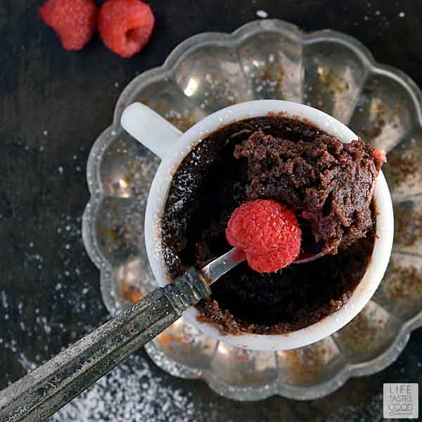 White mug set on a rustic silver fluted saucer filled with gooey chocolate mug cake and topped with fresh raspberries