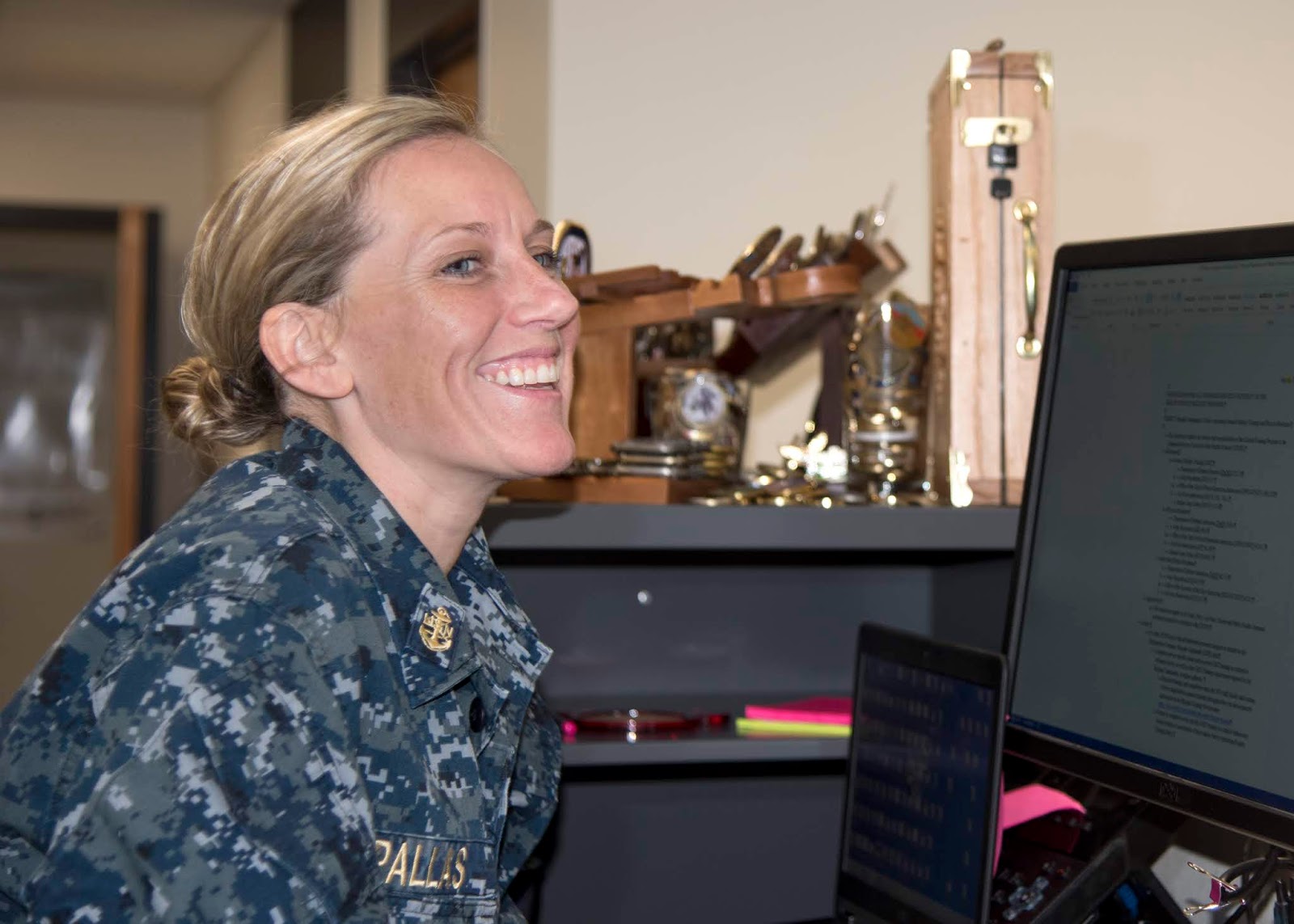 A female in a U.S. Navy uniform is at her desk working. She’s laughing and looking toward her computer.