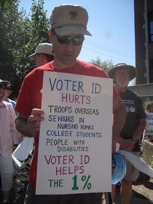 Man holding sign with list of people hurt by requiring voter ID, ending with Helps the 1%