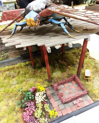 A diorama of a giant insect above a patio and garden bed at a scale model exhibition.