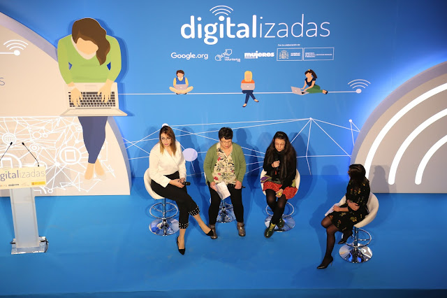 Vista aérea de 4 mujeres en un escenario con un cartel que dice Digitalizadas detrás.
