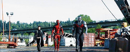 No gif: Míssil Adolescente Megassônico, Deadpool e Colossus caminham em direção à câmera num beco cercado de ferro velho. Ao fundo, uma rodovia onde passam carros como se nada estivesse acontecendo.