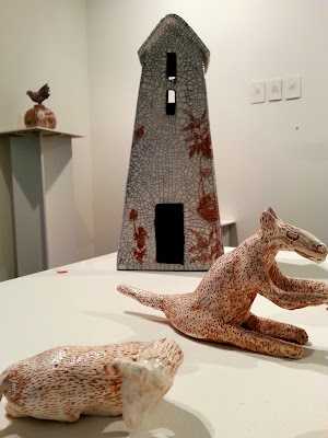 Clay house on display in a gallery. In front of it is a clay kangaroo and sheep.