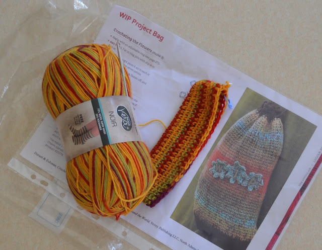 A ball of multicoloured sock yarn (red and yellow mix) with a crochet hook stuck between the label and the yarn.  Attached to the ball of yarn is the beginnings of a striped bag.  All are resting on a photocopied page of the pattern instructions which shows a photo of the finished product.