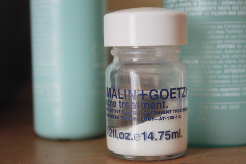 malin + goetz acne treatment sitting on a wooden dressing table, with several Clinique lotions sitting behind it; the formula has separated, so a white powder sits on the bottom, and a clear liquid sits on top.
