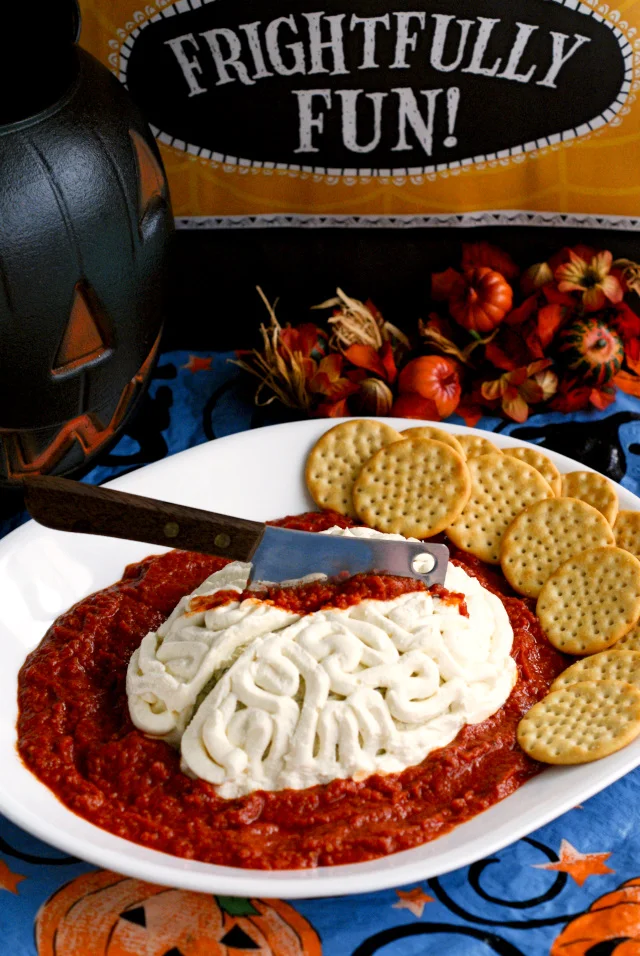 A cheese ball on the shape of a brain with a knife stuck in the top and tomato sauce around the knife and around the cheese ball.